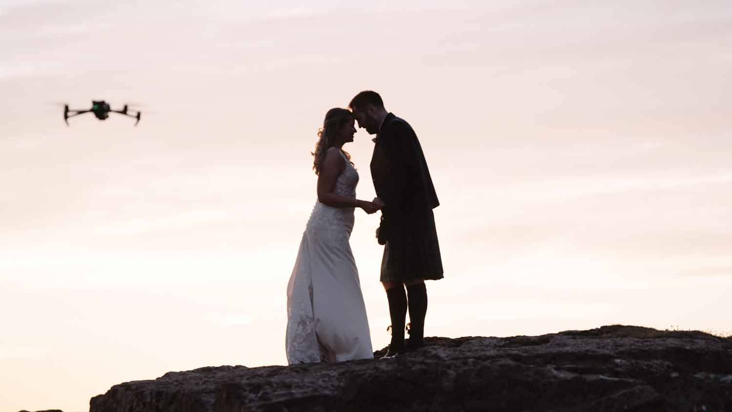 5 Reasons Why a second videographer is a good investment? A bride and Groom posed on a rock with a stunning sunset being filmed by a DJI Mavic 3 in the foreground. 