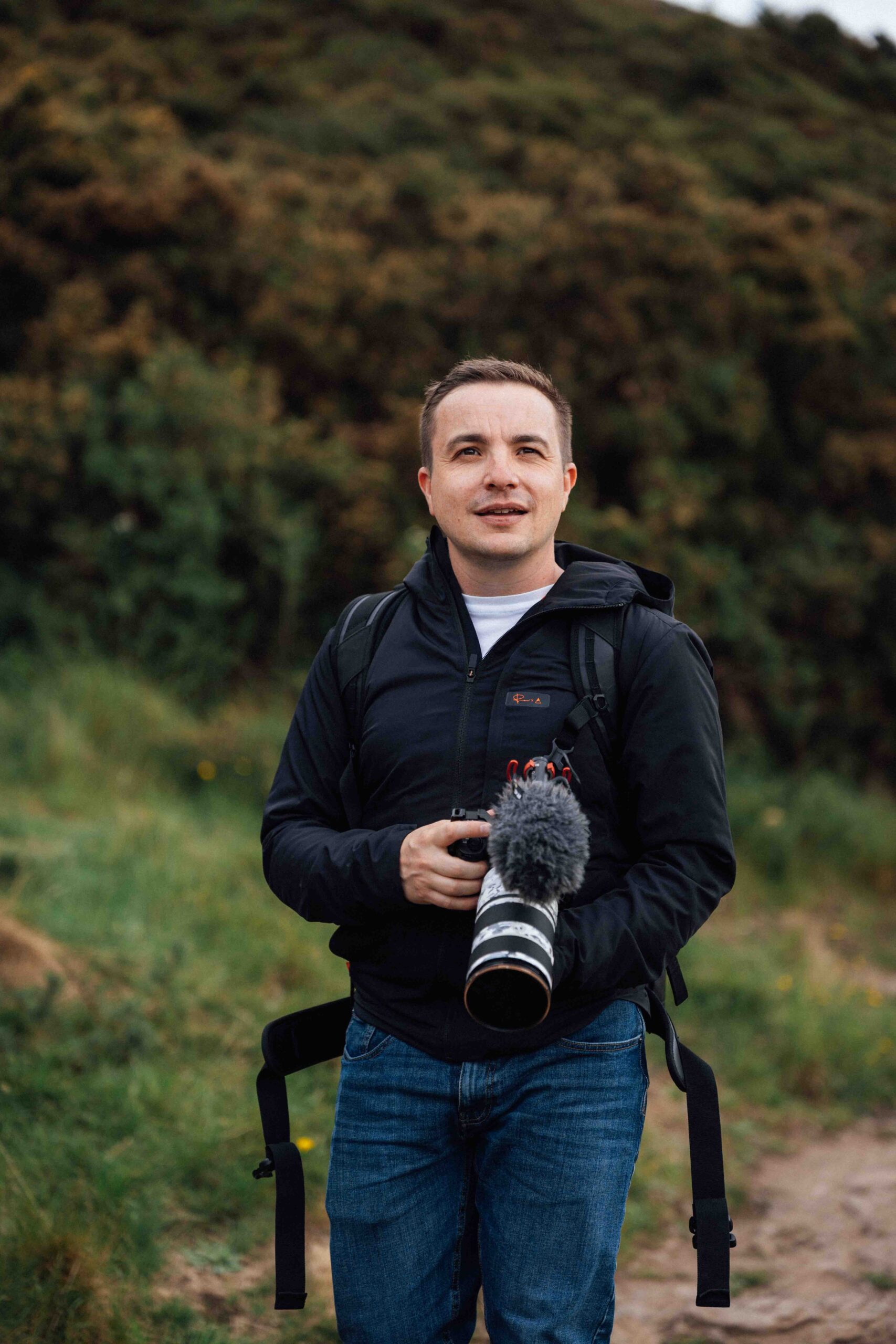 Michael Westcott hiking around arthurs seat holding a sony camera with tele lens looking off into the distance. Shot is showing front side of michael as he looks past the camera.