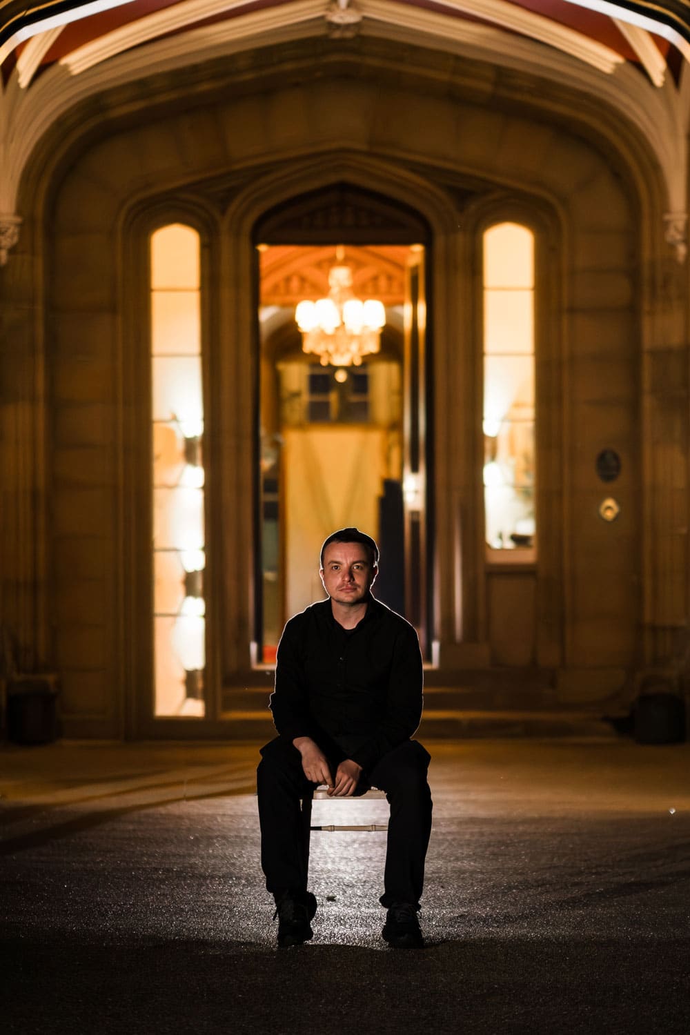 a man dressed in black sits on a chair at night in front of a grand, illuminated building entrance with arched doorways and glowing chandeliers inside, evoking the timeless allure found in documentary wedding videography. copyright michael westcott films