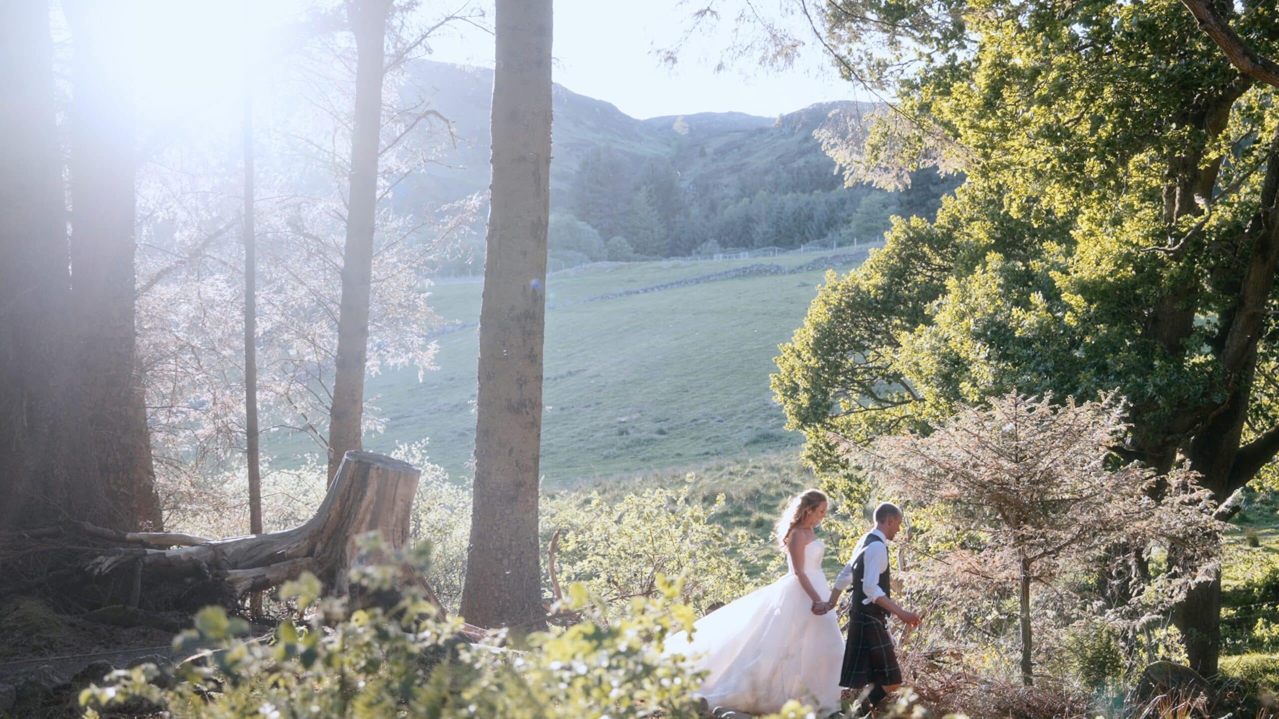 a bride and groom walk hand in hand through a sunlit forest, with green hills surrounding them. captured in a documentary wedding videography style, sunlight streams through the trees, creating a dreamy atmosphere as they stroll together. copyright michael westcott films
