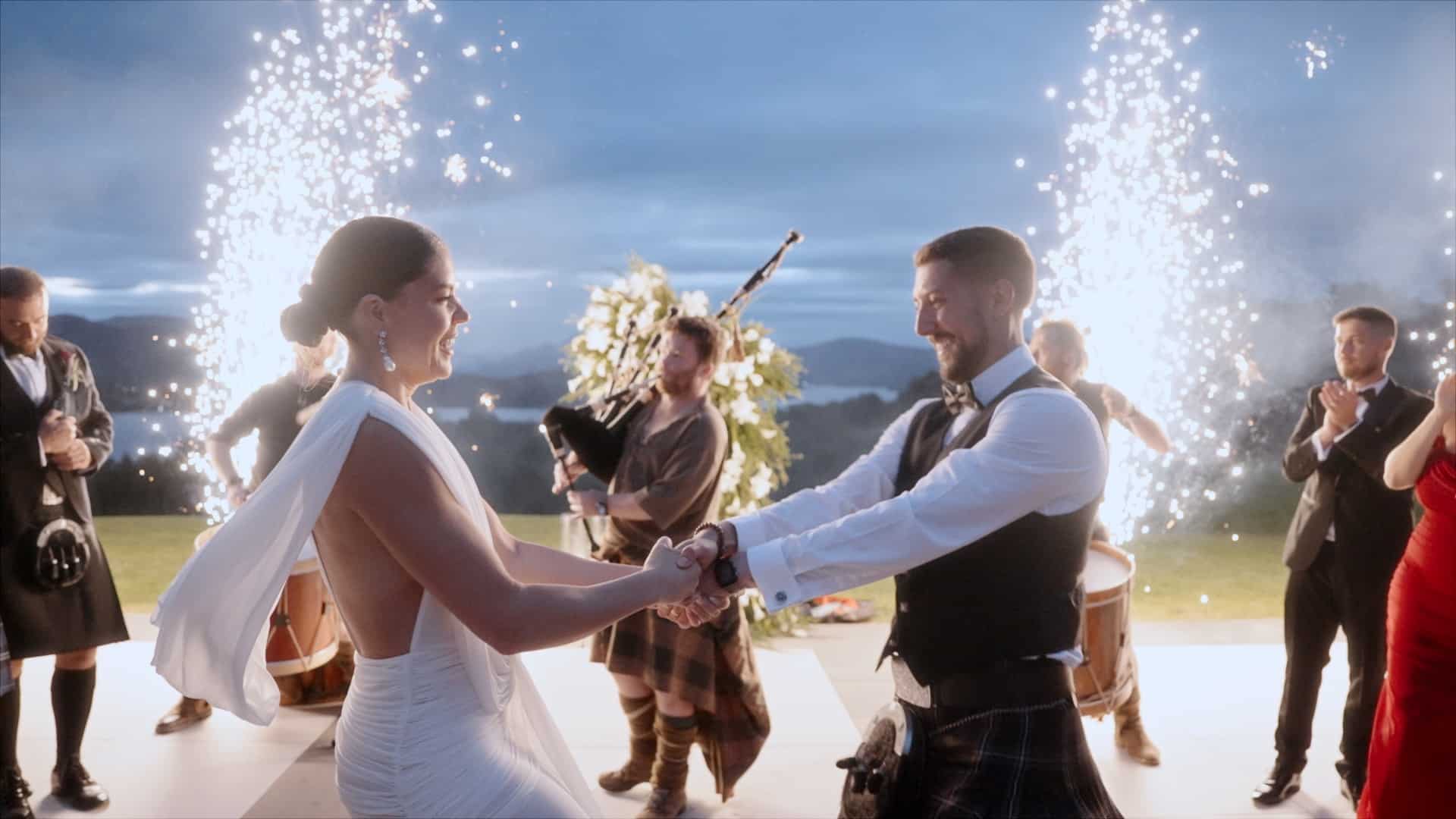 a bride and groom in formal attire dance outdoors at sunset, with bagpipers and drummers in kilts playing nearby and sparklers erupting in the background—capturing the magic of the moment through documentary wedding videography. copyright michael westcott films